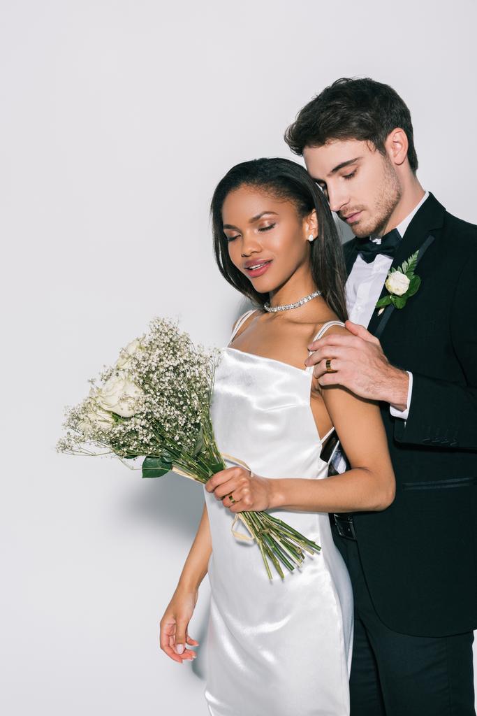 Yakışıklı damat, beyaz arka planda düğün buketi tutan güzel Afrikalı Amerikalı gelinin omzuna dokunuyor. - Fotoğraf, Görsel