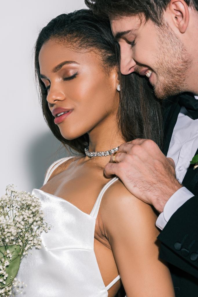 noivo feliz tocando ombro de noiva americana africana bonita segurando buquê de casamento com olhos fechados no fundo branco
 - Foto, Imagem