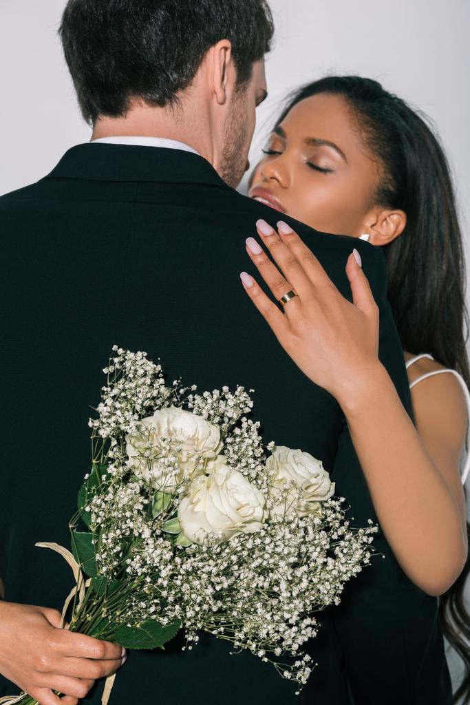 krásný africký americký nevěsta objímání ženich při držení svatební kytice izolované na bílém - Fotografie, Obrázek