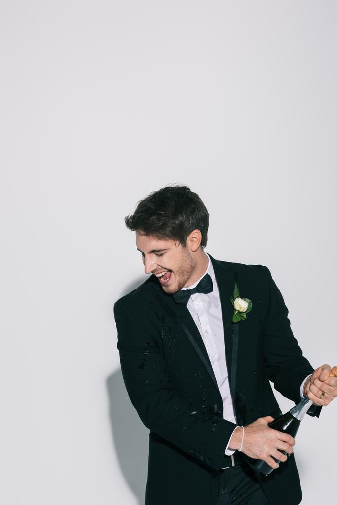 aufgeregter Bräutigam mit geschlossenen Augen öffnet Champagnerflasche auf weißem Hintergrund - Foto, Bild
