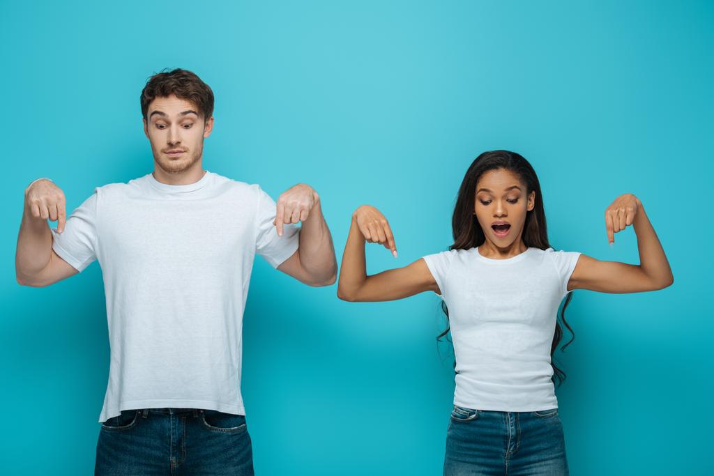 shocked interracial couple pointing with fingers and looking down on blue background  - Photo, Image