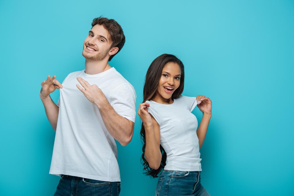 cheerful interracial couple touching white t-shirts on blue background - Photo, Image