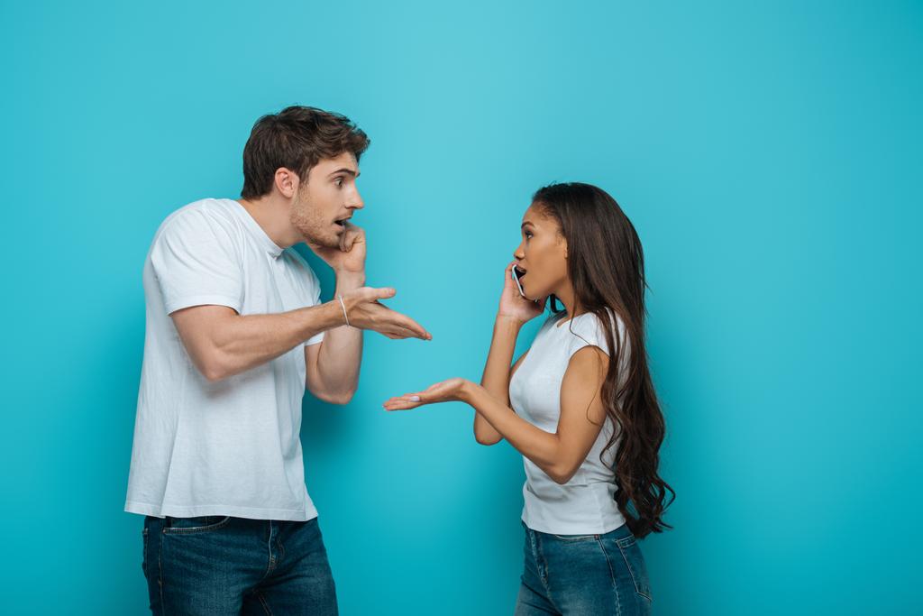 Seitenansicht eines jungen gemischtrassigen Paares, das sich streitet, während es auf Smartphones auf blauem Hintergrund spricht - Foto, Bild