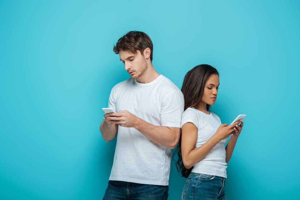 young interracial couple chatting on smartphones while standing back to back on blue background - Photo, Image