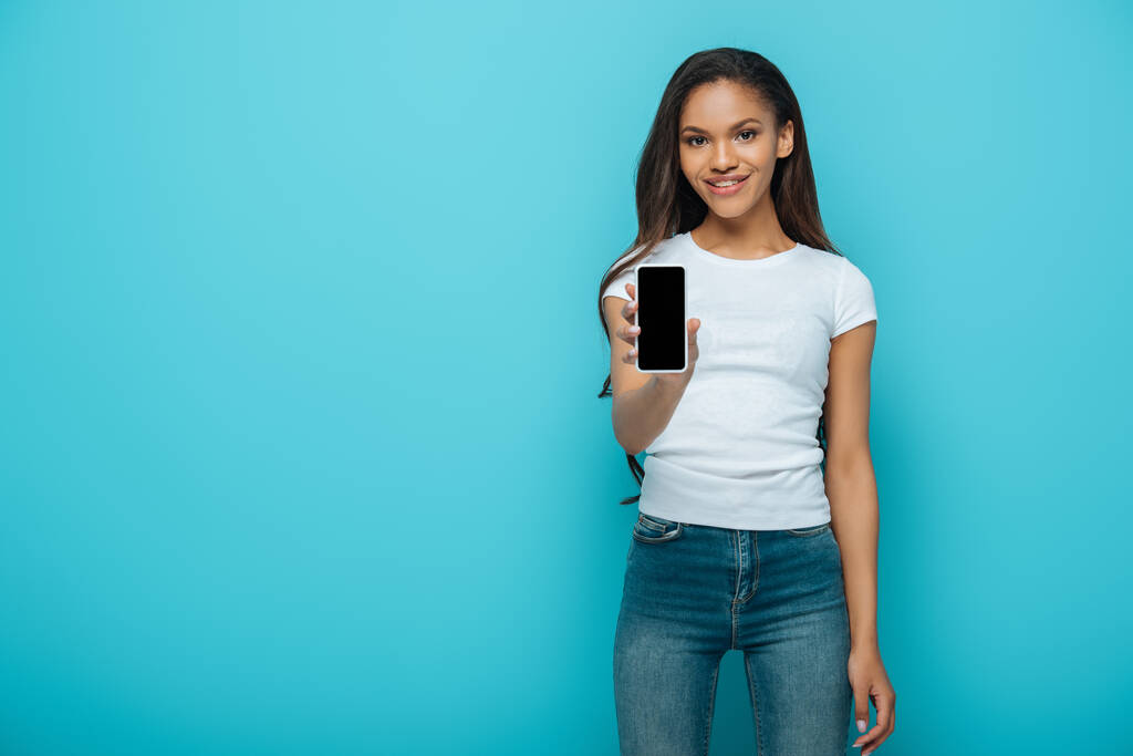 hymyilevä afrikkalainen amerikkalainen tyttö osoittaa älypuhelin tyhjä näyttö eristetty sininen
 - Valokuva, kuva