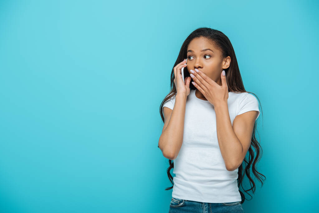 impactó a chica afroamericana cubriendo la boca con la mano mientras hablaba en un teléfono inteligente aislado en azul
 - Foto, Imagen