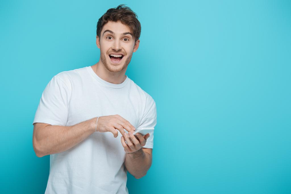 vzrušený chlap při pohledu na kameru při chatování na smartphone na modrém pozadí - Fotografie, Obrázek