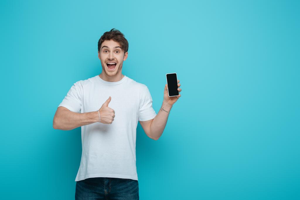 jovem animado mostrando polegar para cima enquanto segurando smartphone com tela em branco no fundo azul
 - Foto, Imagem