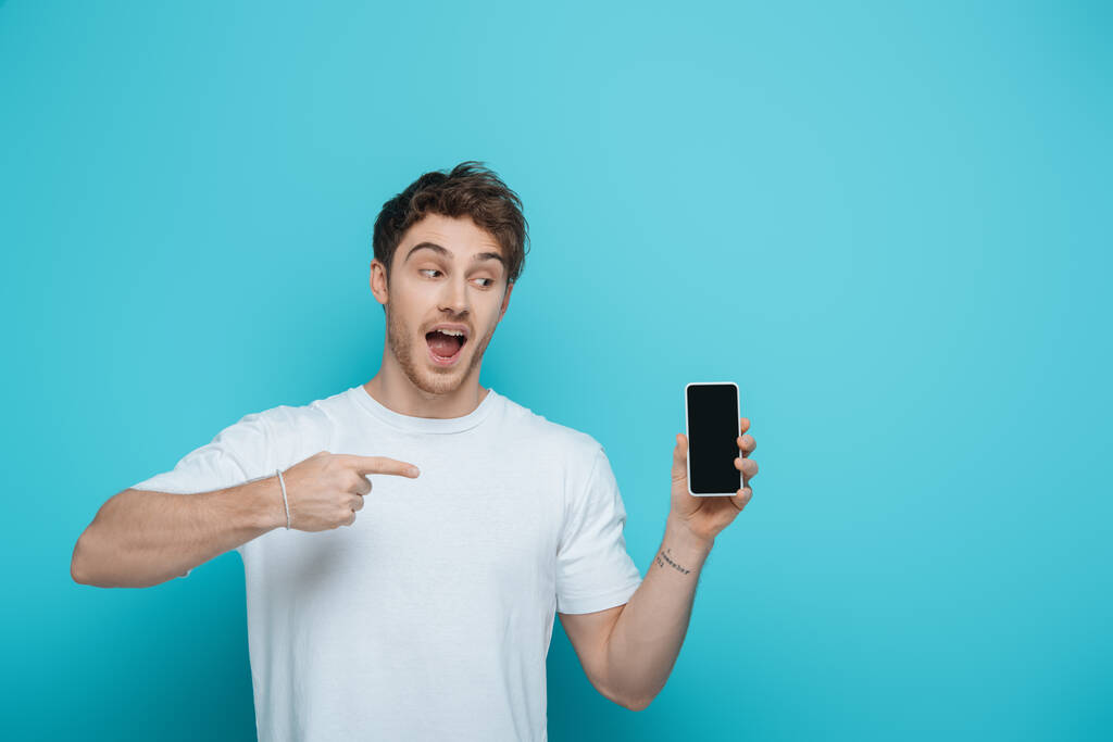 興奮した男は青の背景に空白の画面を持つスマートフォンで指を指して - 写真・画像