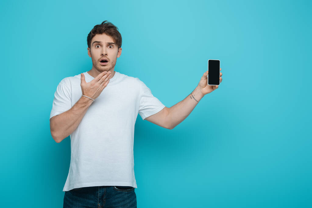 wstrząśnięty facet trzymając rękę blisko twarzy podczas wyświetlania smartfona z pustym ekranem na niebieskim tle - Zdjęcie, obraz
