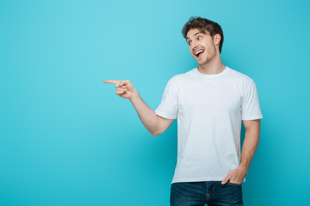 smiling guy holding hand in pocket, looking away and pointing with finger on blue background - Photo, Image
