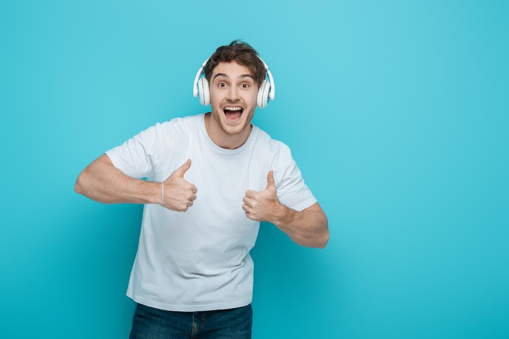 excited young man in wireless headphones showing thumbs up while looking at camera on blue background - Photo, Image