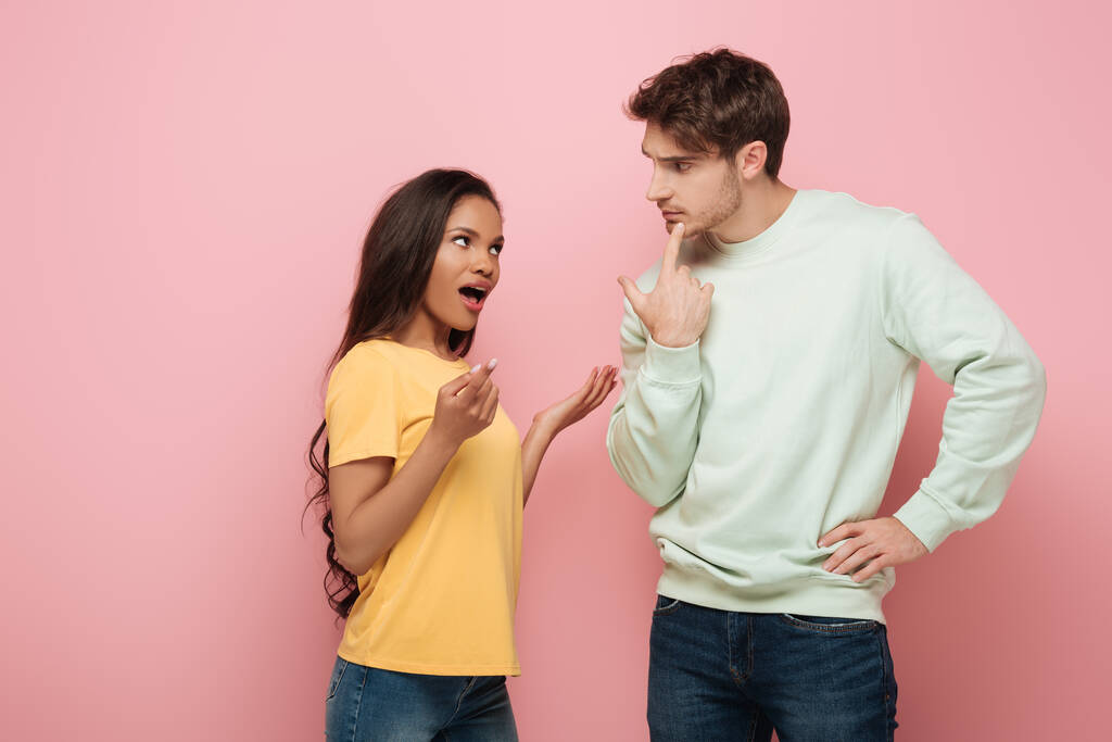 serious guy touching chin while looking at african american girlfriend quarreling on pink background - Photo, Image