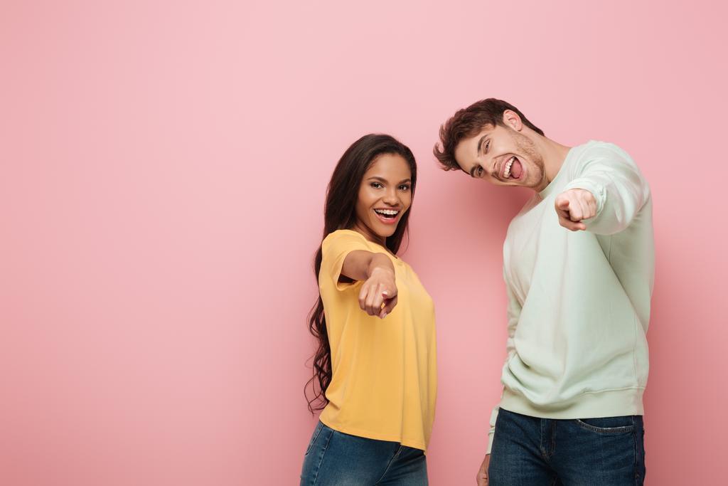 excited interracial couple pointing with fingers at camera on pink background - Photo, Image