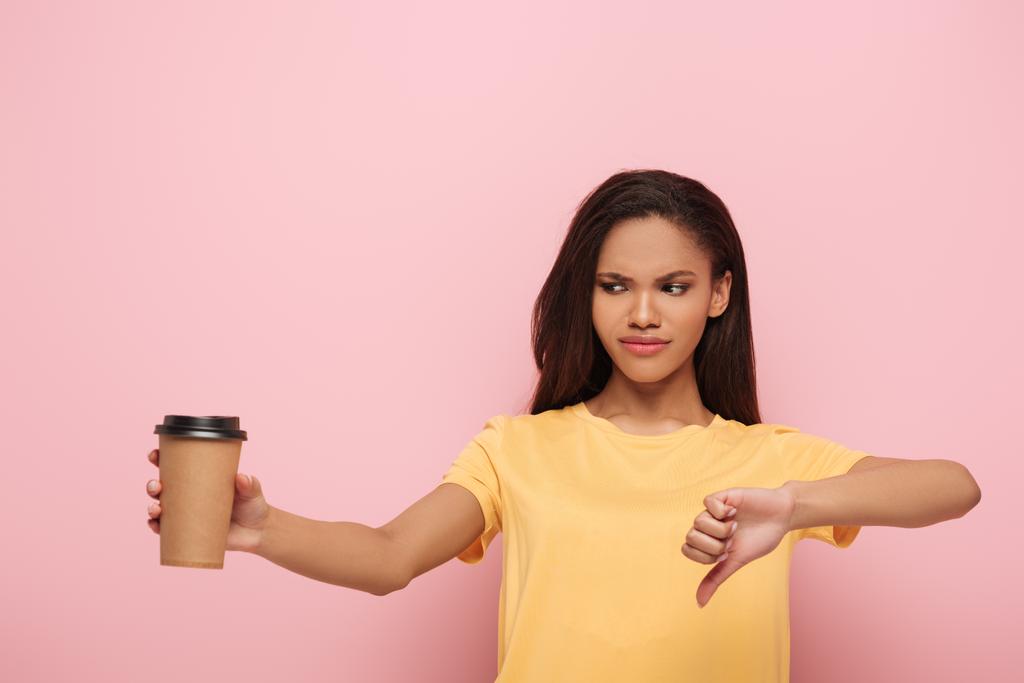 displeased african american girl showing thumb down while holding coffee to go on pink background - Photo, Image