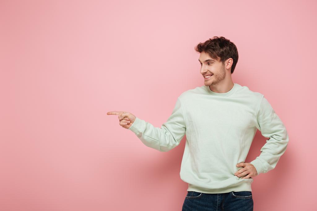 fröhlicher junger Mann, der wegschaut und mit dem Finger zeigt, während er die Hand an der Hüfte auf rosa Hintergrund hält - Foto, Bild