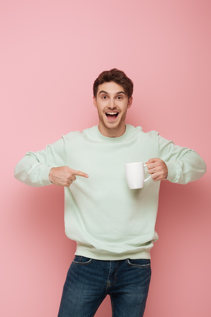 excited guy pointing with finger at white cup while looking at camera on pink background - Photo, Image