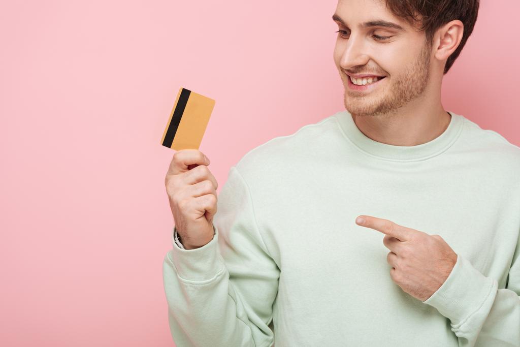 šťastný mladý muž ukazuje prstem na kreditní kartu na růžovém pozadí - Fotografie, Obrázek