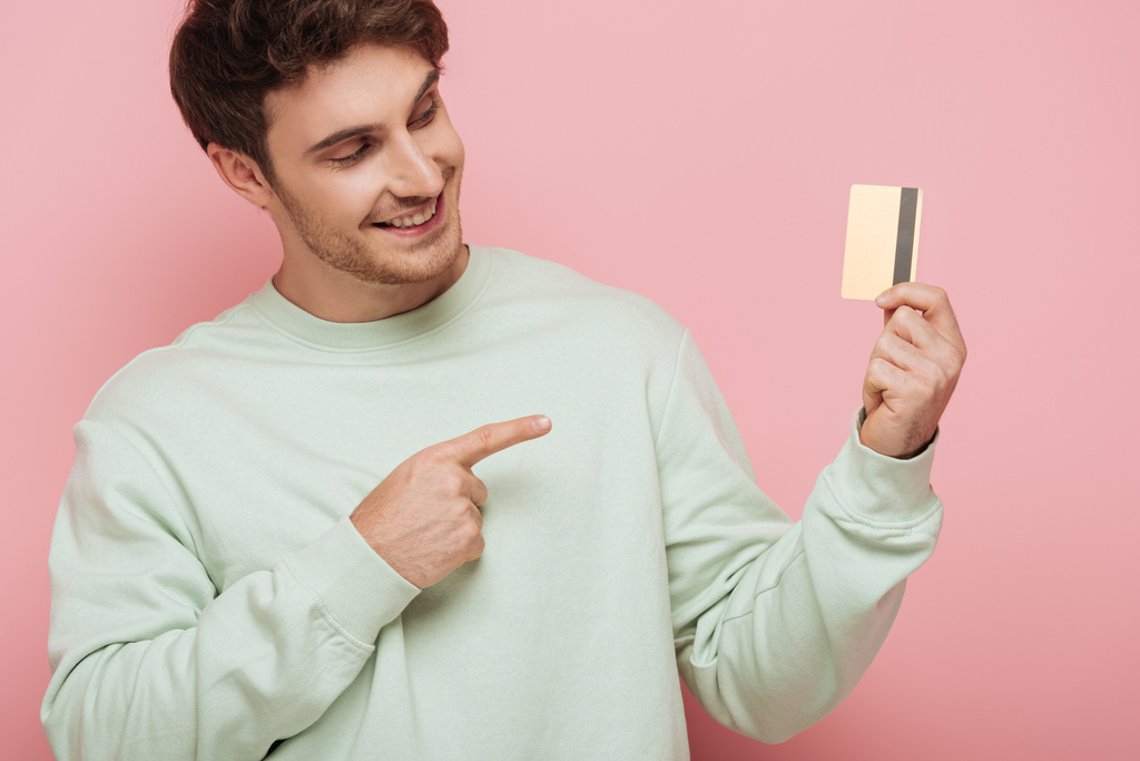 sonriente joven apuntando con el dedo a la tarjeta de crédito sobre fondo rosa
 - Foto, Imagen