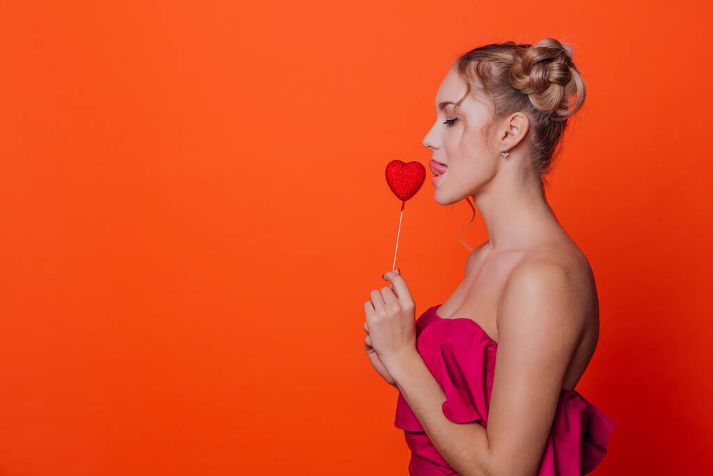 Το κορίτσι με το κόκκινο φόρεμα σε πορτοκαλί φόντο στο στούντιο. Ξανθιά κοπέλα με δύο κόμπους στα μαλλιά που κρατάει κόκκινη καρδιά στο κοντάρι κοντά στα χείλη. Αντιγραφή χώρου. - Φωτογραφία, εικόνα