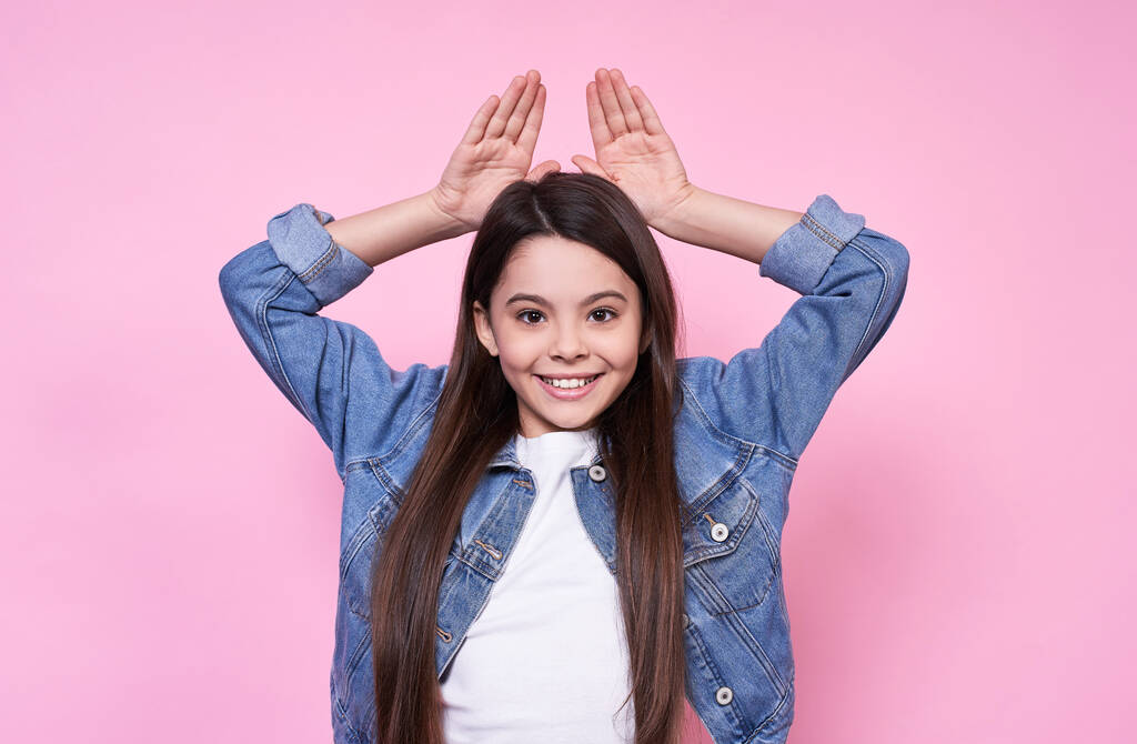 Cute young funny emotional Caucasian girl child long-haired brunette. Laugh and smile and holds hands behind his head while mimicking a rabbit. In white t-shirt jeans jacket on a pink background. - Photo, Image