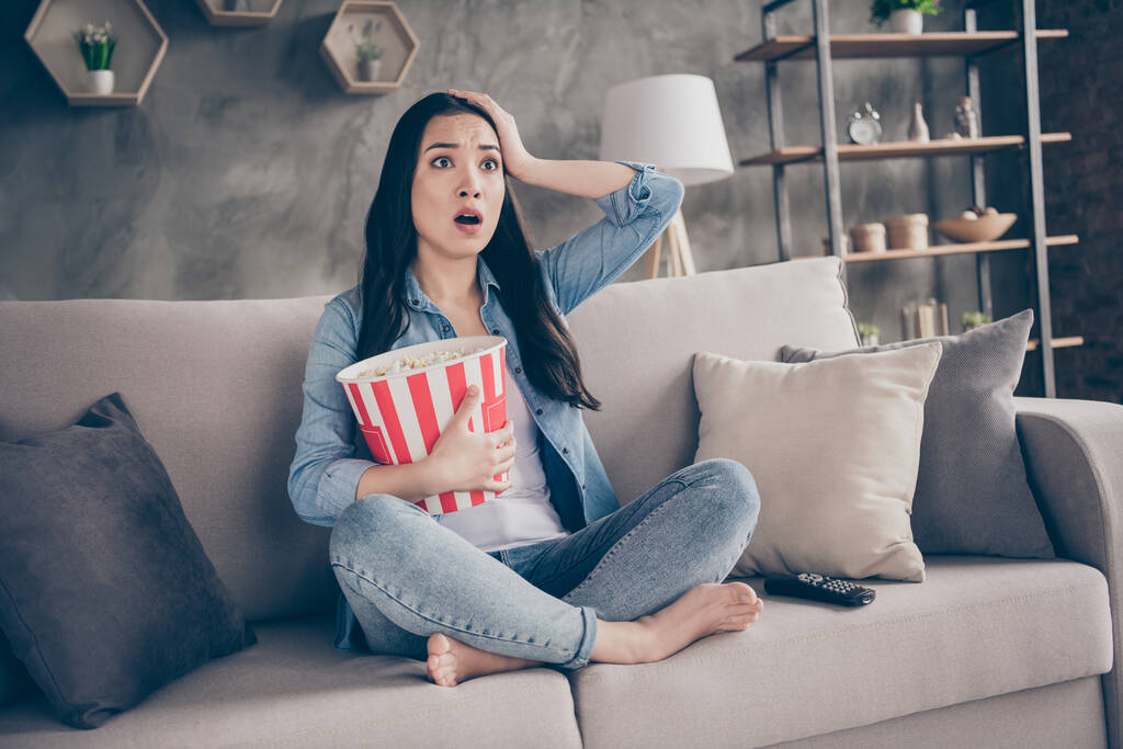 Full size photo of scared girl sit divan legs cross folded barefoot watch horor film hold big pop corn box impressed touch hand head in apartment room - Fotografie, Obrázek