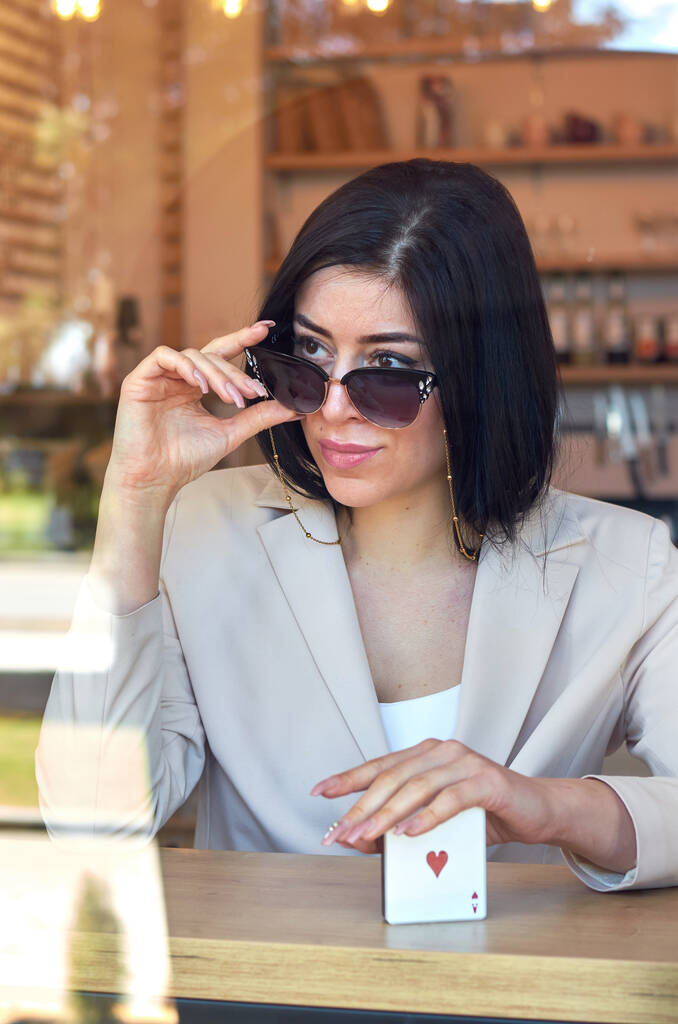 Fotografie módní, moderní, stylové ženy, která drží sluneční brýle se zlatým řetízkem přes skleněnou kavárnu. - Fotografie, Obrázek