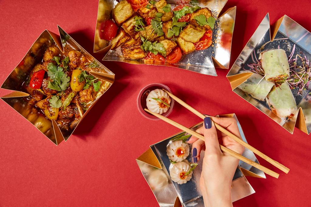 κορυφαία άποψη της γυναίκας κρατώντας chopsticks με ψωμάκι στον ατμό κοντά στο δοχείο με σάλτσα και παρασκευασμένο κινέζικο φαγητό σε κουτιά takeaway στο κόκκινο - Φωτογραφία, εικόνα