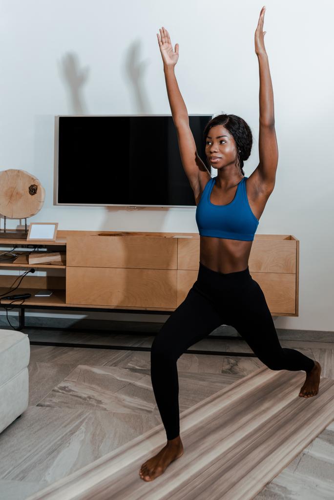 Femme sportive afro-américaine debout dans la pose de haute fente avec les mains dans l'air et souriant sur tapis de yoga dans le salon
 - Photo, image