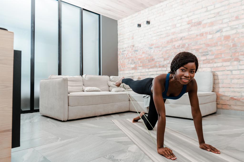 Sportiva afroamericana a quattro zampe che pratica yoga con banda di resistenza, sorridente e guardando la macchina fotografica in soggiorno
 - Foto, immagini