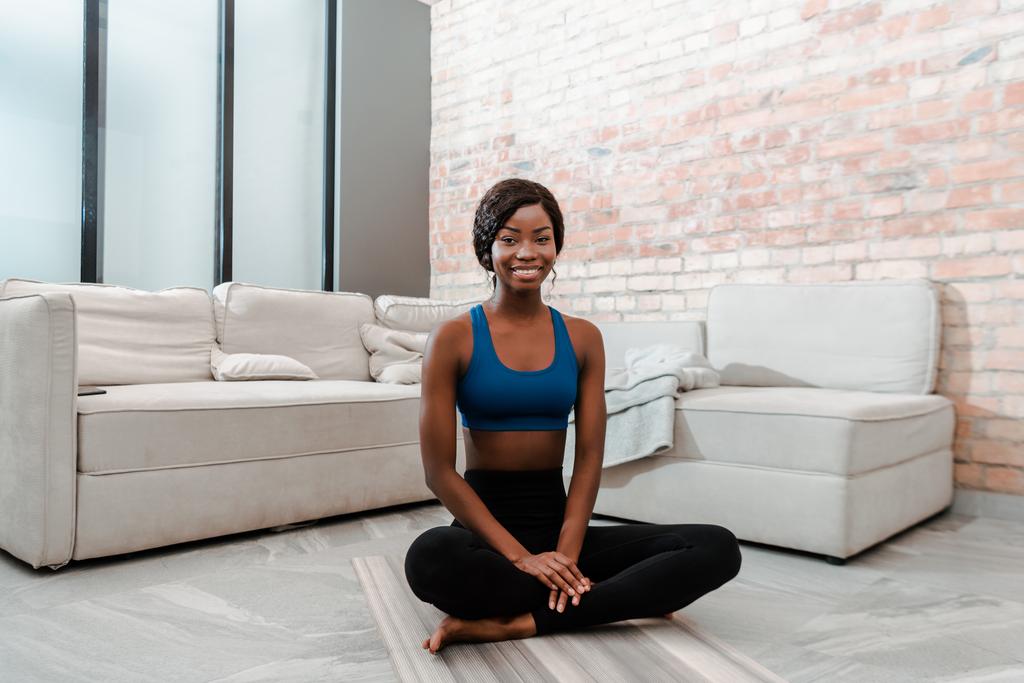 Африканська спортсменка з Америки посміхається, дивиться на камеру і сидить у позі лотоса на йога маті у вітальні. - Фото, зображення