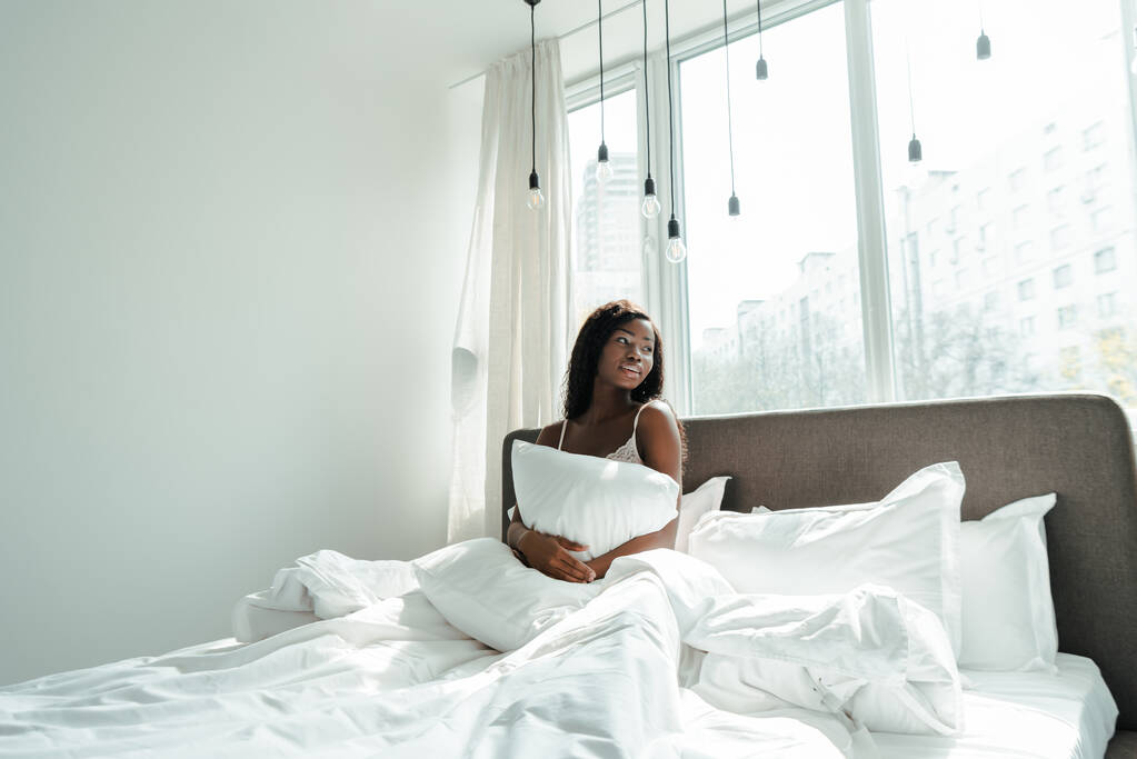 African american woman holding pillow and looking away on bed in bedroom - Photo, Image