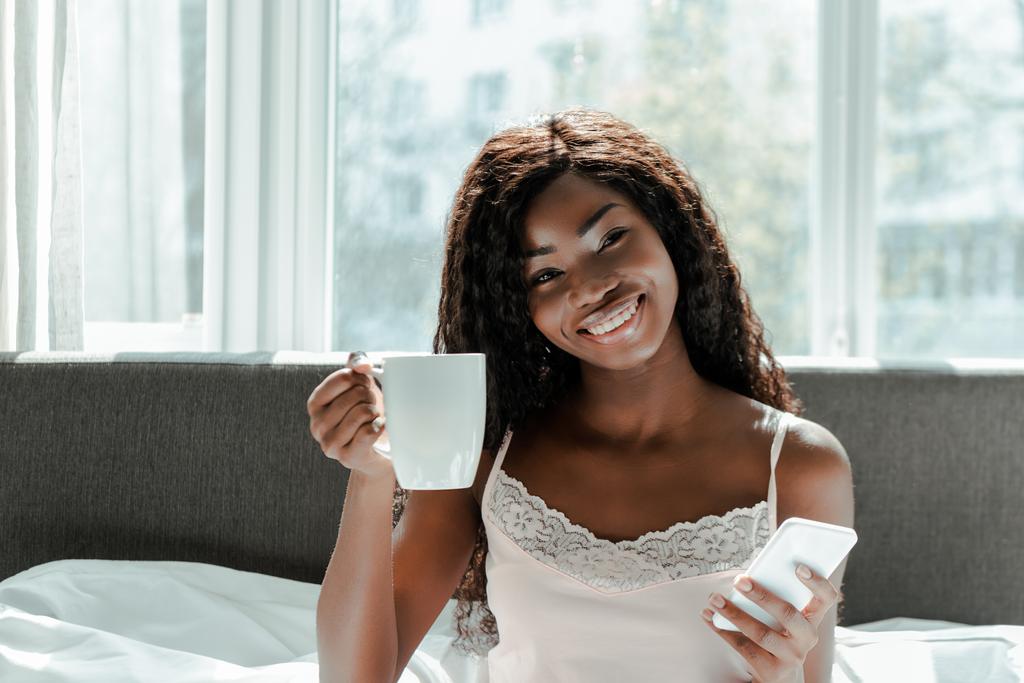 Mujer afroamericana con teléfono inteligente sonriendo, mostrando taza de té y mirando a la cámara en la cama en el dormitorio
 - Foto, imagen