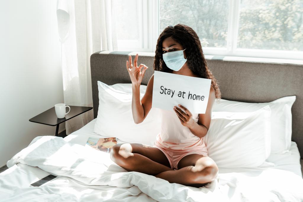 African american woman in medical mask with crossed legs showing okay sign and placard with stay at home lettering on bed in bedroom - Photo, Image