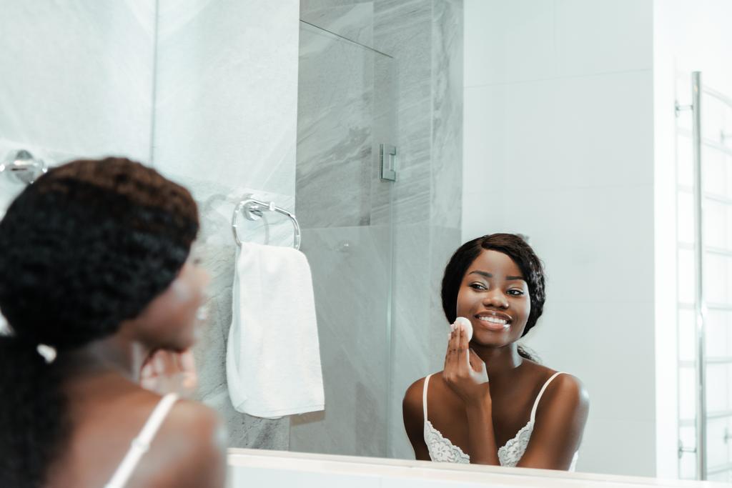 Африканська американка витирає обличчя бавовняною подушкою, дивиться на дзеркало і посміхається у ванній кімнаті. - Фото, зображення