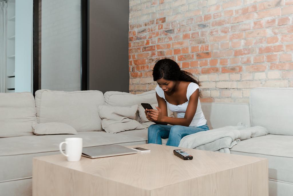 Afrikai amerikai nő digitális tabletta kanapén közel dohányzóasztal laptop, okostelefon, távirányító és csésze tea a nappaliban - Fotó, kép