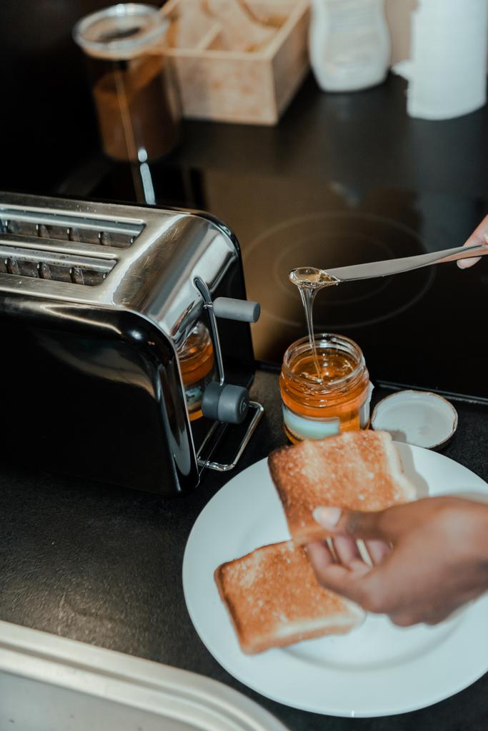 Розбитий вигляд на африканську американську жінку, яка готує тост з медом біля тостера на кухні. - Фото, зображення