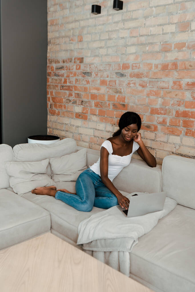 African american freelancer using laptop on sofa in living room - Photo, Image