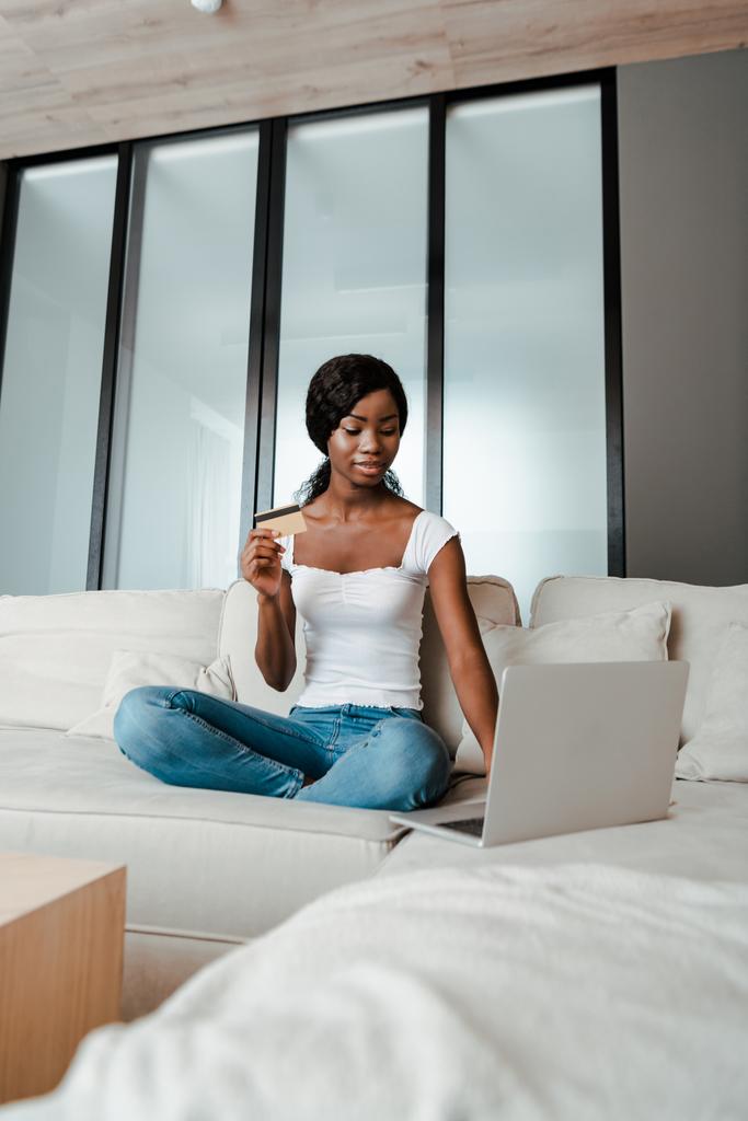 Африканська американка дивиться на ноутбук і показує кредитну картку на дивані у вітальні. - Фото, зображення
