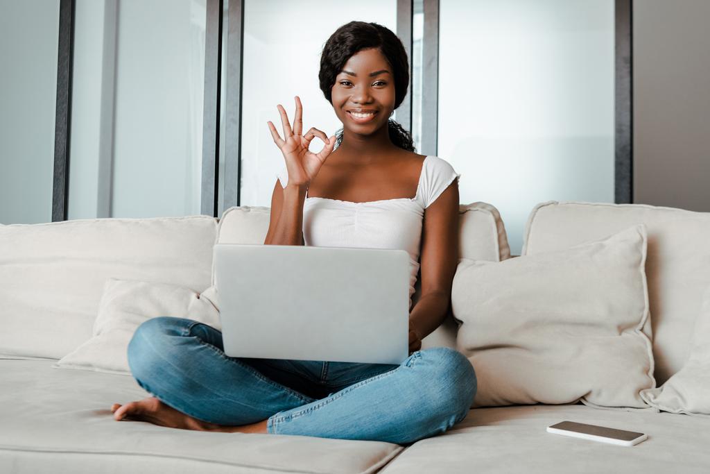 Afrikkalainen amerikkalainen freelancer ristissä jalat ja kannettava tietokone hymyilee ja näyttää kunnossa merkki lähellä älypuhelin sohvalla olohuoneessa
 - Valokuva, kuva