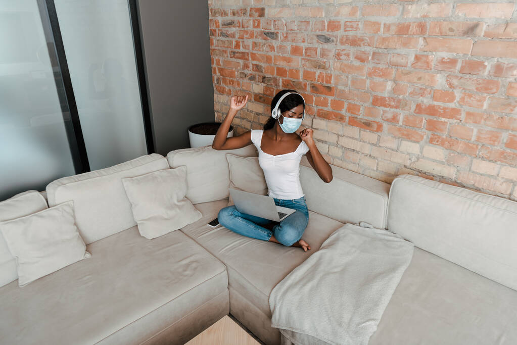 Високий кут зору афроамериканського фрілансера в медичній масці та навушники, що слухають музику з ноутбуком на дивані у вітальні
 - Фото, зображення