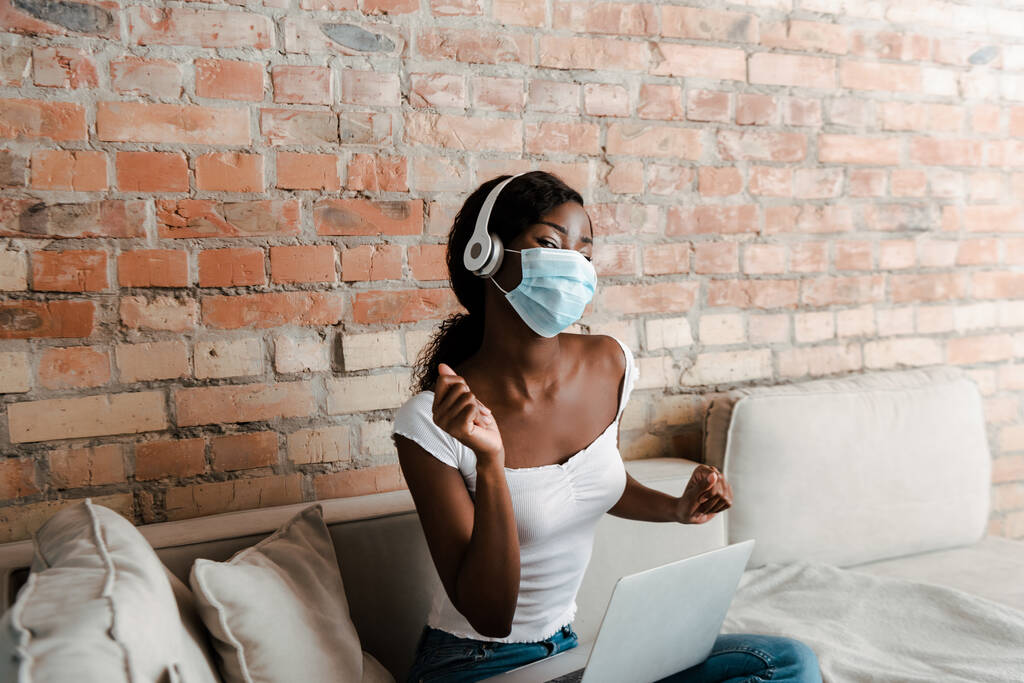 Freelancer afroamericano con máscara médica y portátil escuchando música en auriculares cerca de la pared de ladrillo en la sala de estar
 - Foto, Imagen