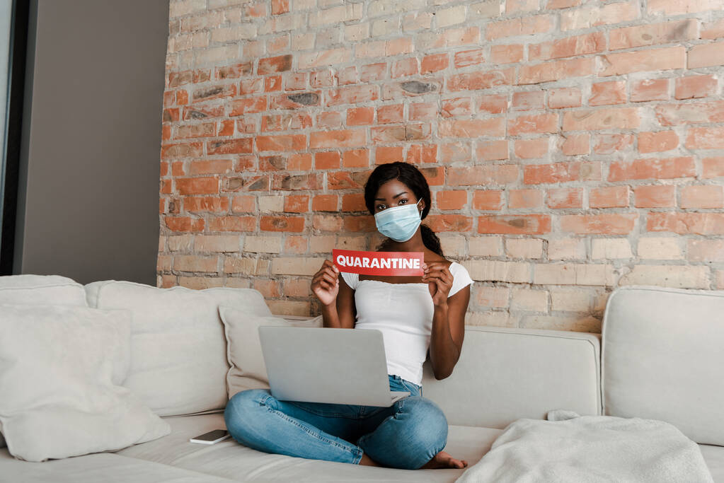 Afroamerikanischer Freiberufler in medizinischer Maske mit überkreuzten Beinen und Laptop zeigt Karte mit Quarantäne-Schriftzug auf Sofa im Wohnzimmer - Foto, Bild