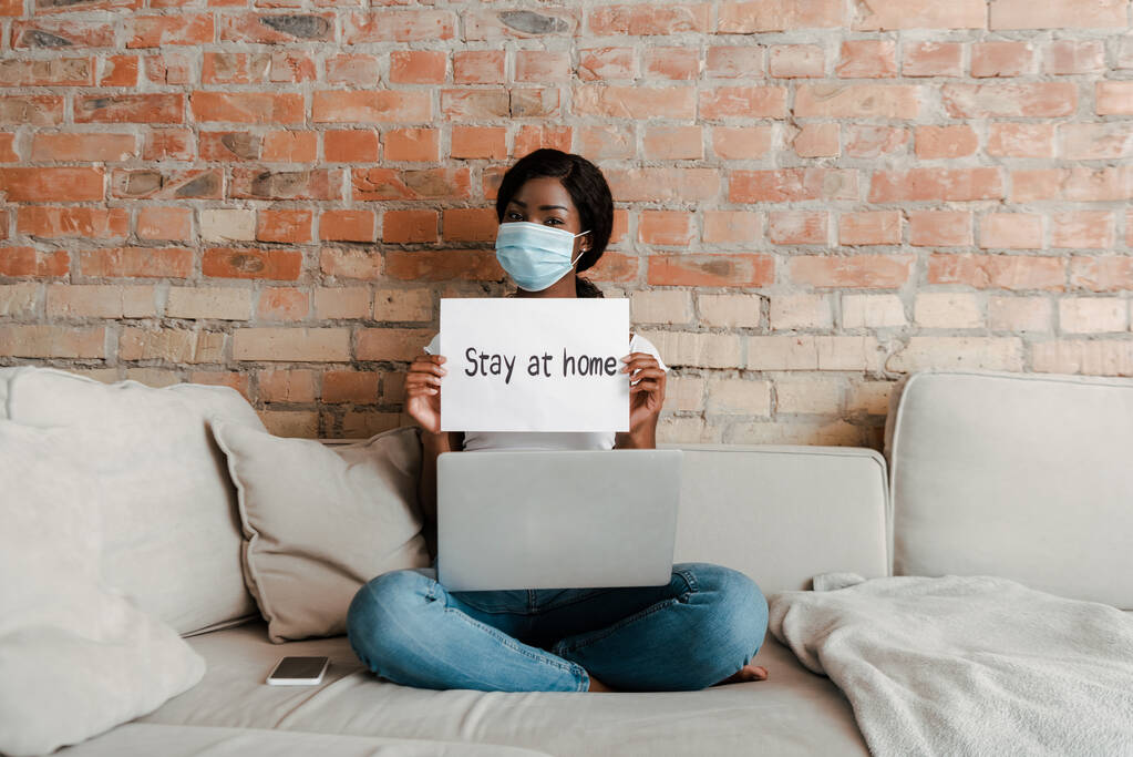 African american freelancer in medical mask with laptop near smartphone showing placard with stay at home lettering on sofa in living room - Photo, Image