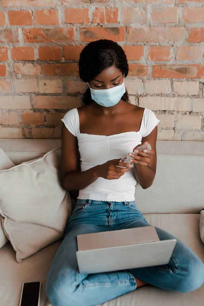 Afro-Américain pigiste en masque médical avec ordinateur portable près du smartphone tenant désinfectant pour les mains sur le canapé dans le salon
 - Photo, image