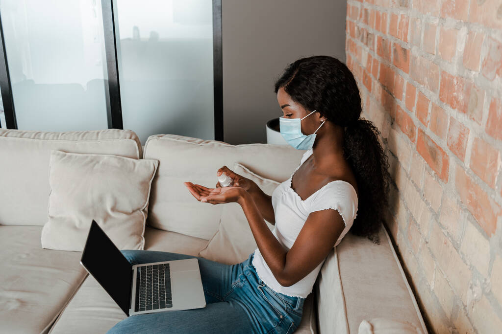 Wysoki kąt widzenia afrykański amerykański freelancer w masce medycznej z laptopem za pomocą środka do dezynfekcji dłoni na kanapie w salonie - Zdjęcie, obraz