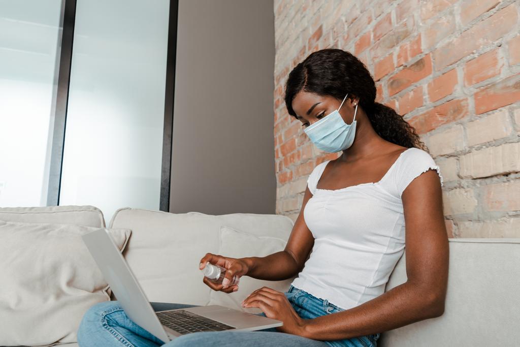 Indépendant afro-américain dans un masque médical désinfectant ordinateur portable avec désinfectant pour les mains sur canapé dans le salon
 - Photo, image