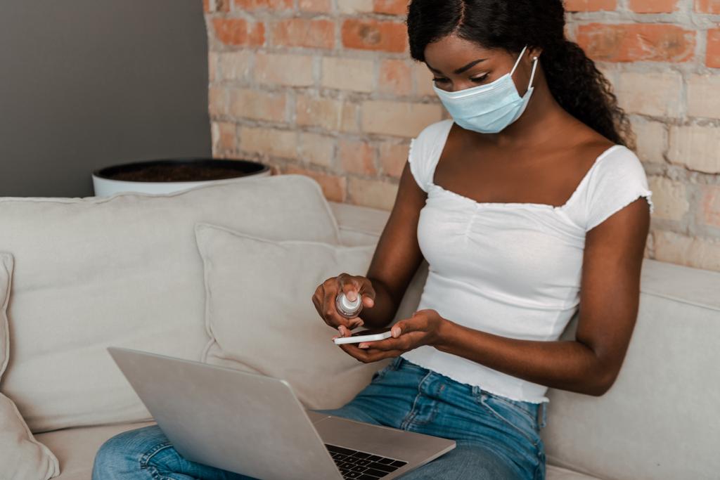 Αφροαμερικανός ελεύθερος επαγγελματίας σε ιατρική μάσκα με φορητό υπολογιστή απολύμανση smartphone με απολυμαντικό χεριών σε καναπέ στο σαλόνι - Φωτογραφία, εικόνα