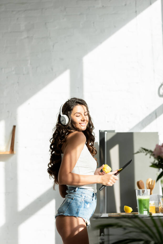Vista lateral da mulher sexy em fones de ouvido sorrindo enquanto segurando pedaço de limão e faca na cozinha
  - Foto, Imagem