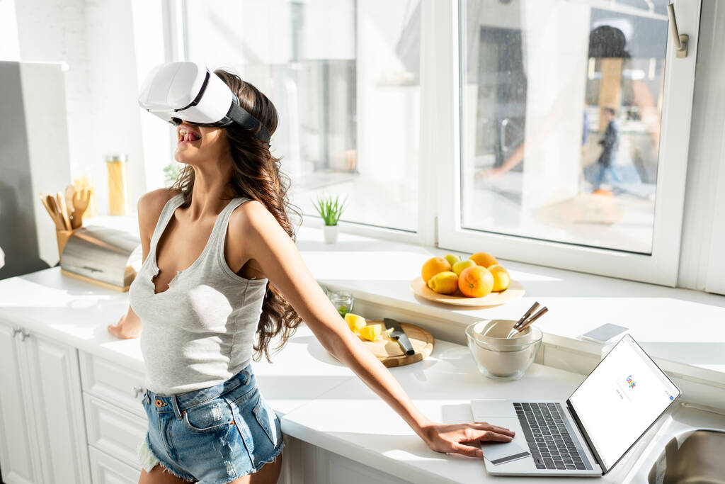 KYIV, UKRAINE - MARCH 13, 2020: Young woman using vr headset near laptop with google website and credit card in kitchen  - Photo, Image